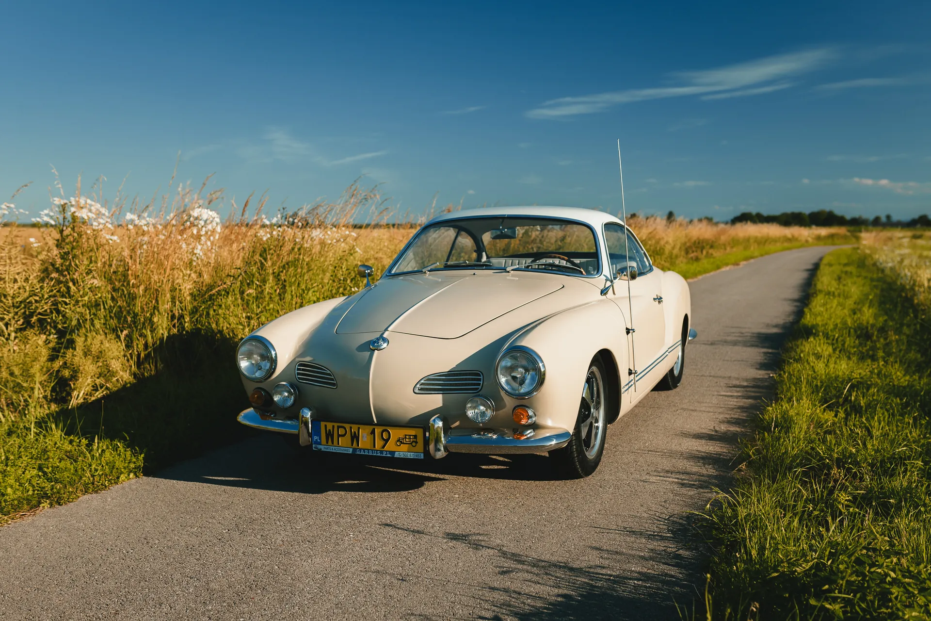 Kremowy Volkswagen Karmann Ghia na wąskiej drodze wśród pól w słoneczny dzień