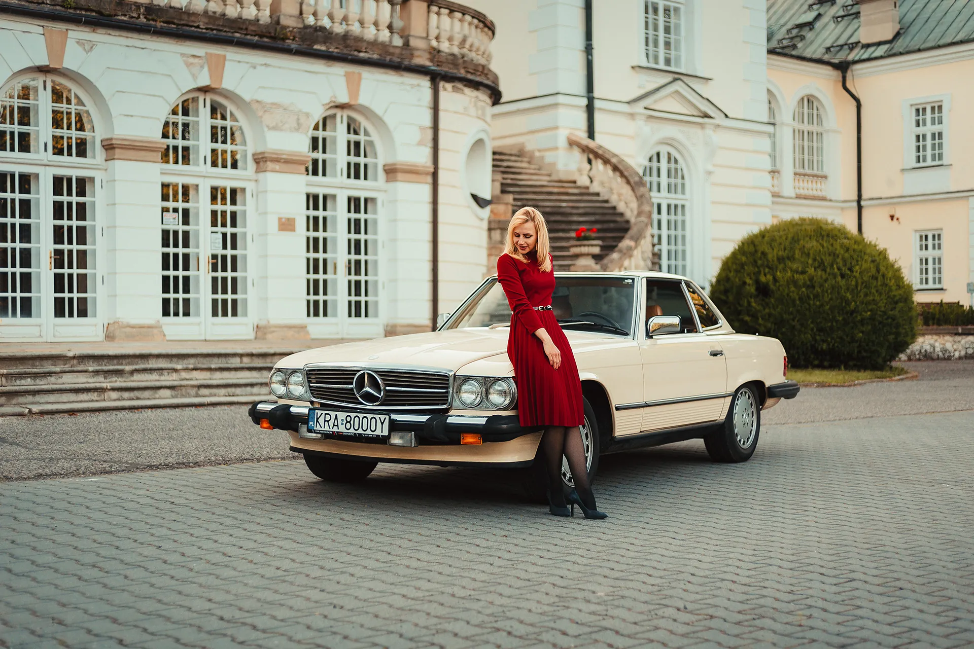 Kobieta w czerwonej sukience przy kremowym klasycznym Mercedesie przed pałacem